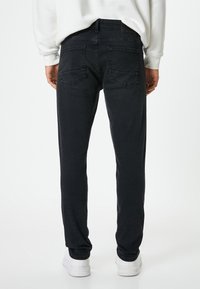 Jeans de denim negro con corteSlim, que cuentan con dos bolsillos traseros y una textura suave. Combinados con una zapatilla blanca y detalles de costura visibles.