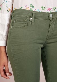 Jeans verde oliva con un diseño ajustado, que cuentan con un bolsillo frontal, un cierre de botón metálico y una tela suave y texturizada.
