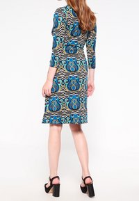 Fitted dress with three-quarter sleeves, colorful circular patterns on a striped background, black accentuated hem, paired with chunky heeled sandals.