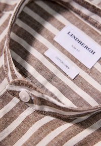 Cuello de camisa de algodón y lino con un patrón de rayas marrones y blancas; cuenta con un cierre de botón y etiquetas que indican la marca y el material.