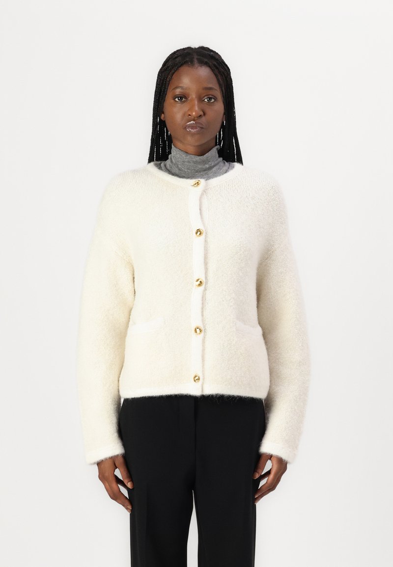 Cardigan blanc et duveteux avec un col rond, fermeture à boutons sur le devant et deux poches ; présente des boutons dorés et une texture douce.
