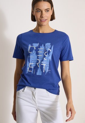 Femme portant un t-shirt graphique bleu avec des coups de pinceau abstraits blancs et bleu clair et des lettres, associé à un pantalon blanc.