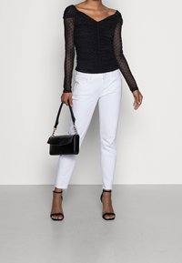 Top nero a spalle scoperte con maniche lunghe trasparenti e texture a pois, abbinato a jeans bianchi slim-fit e una piccola borsa nera.