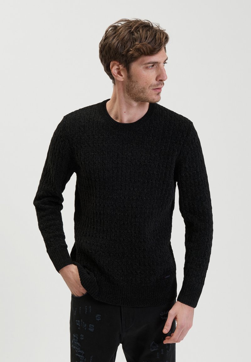 Maglione nero lavorato a maglia con un motivo texturizzato, scollatura tonda e polsini a coste. Indossato con pantaloni neri con un design sottile.