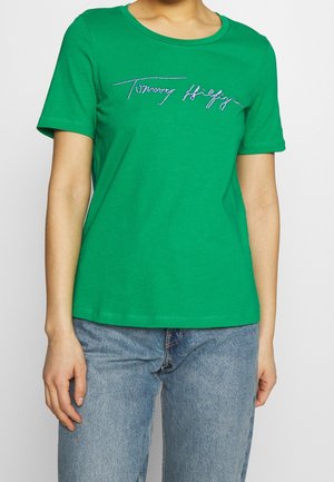 Personne portant un t-shirt vert à manches courtes avec le logo "Tommy Hilfiger" en strass et un jean bleu clair, debout devant un fond uni.