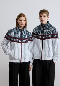 Λευκά σακάκια με κυματιστό σχέδιο σε μπλε και πράσινο, με διακριτικές μπορντό ρίγες, που διαθέτουν φερμουάρ μπροστά και ελαστικά μανσέτια.