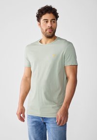 Camiseta de algodón verde claro con cuello redondo, mangas cortas y un pequeño logotipo amarillo bordado en el pecho. Corte casual.