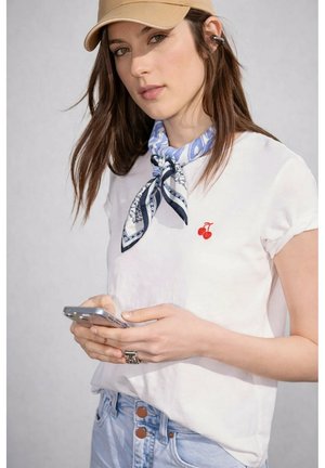 Jeune femme portant une casquette beige, un t-shirt blanc avec un logo cerise, un foulard bleu à motifs autour du cou et un jean, tenant un smartphone à deux mains.
