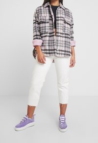 Vrouw draagt een roze en zwarte geruite button-up jas, zwarte cropped top, witte cropped jeans en paarse suède sneakers met witte zolen.