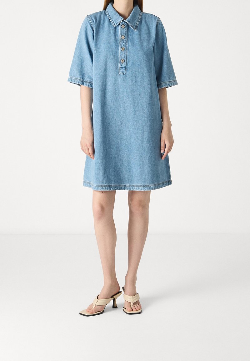 Femme portant une robe en jean bleu clair à manches courtes avec des boutons et des sandales beiges à talons, debout sur un fond blanc.
