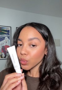 Hvit tube med spiss tupp, merket "LH cosmetics," holdt i en hånd. Modell med langt, bølgete mørkt hår og naturlig makeup, poserer.