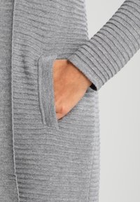 Cardigan grigio a maglia con texture a coste e tasca. Tessuto morbido e maniche lunghe. Il design è aperto sul davanti per una facile vestibilità.