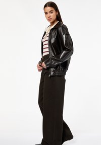 Giovane donna che indossa una giacca di pelle nera sopra una polo a righe e pantaloni a vita alta e larghi scuri, in piedi contro uno sfondo bianco.