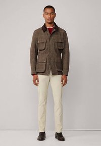 Veste marron à motif à carreaux avec poches à rabat boutonnées, col en velours côtelé, portée sur une chemise rouge, associée à un pantalon crème clair et des chaussures foncées.