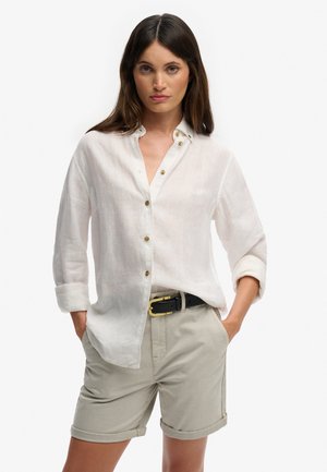 Kobieta ubrana w białą, zapinaną na guziki koszulę z długim rękawem, z podwiniętymi mankietami, oraz beżowe szorty z czarnym paskiem, stoi z rękami w kieszeniach.