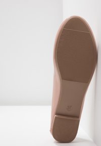 Semelle d'une chaussure couleur nude avec grip texturé et taille 37 marquée, positionnée debout contre une surface d'angle blanche.