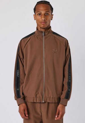 Giacca sportiva marrone con zip e colletto alto, strisce nere decorative sulle spalle e polsini elasticizzati. Presenta il logo sul petto.