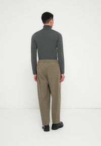 Mand står med ryggen til iført en mørkegrå turtleneck, løse olivengrønne bukser med en baglomme og sorte sko foran en ensfarvet hvid baggrund.