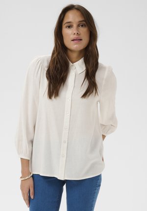 Moteris su ilgais rudos spalvos plaukais, vilkinti baltą palaidinę su pūstomis rankovėmis ir mėlynomis džinsais, stovinti priešais paprastą šviesų foną.