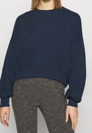 Kobieta ubrana w granatowy krótki sweter z długim rękawem i teksturowane szare legginsy z wysokim stanem, stoi z rozluźnionymi ramionami.