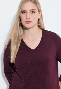 Bordeauxfarbendes Langarmshirt mit V-Ausschnitt, das ein strukturiertes Design mit glitzernden Akzenten aufweist. Weicher Stoff, lockere Passform, minimale Details.