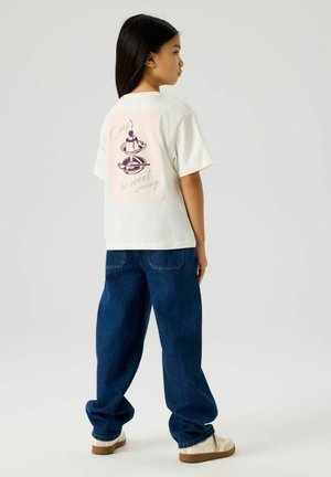 Jente med langt svart hår iført en hvit T-skjorte med desserttrykk og tekst, løse blå jeans og hvite joggesko, sett fra siden.