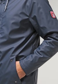 Marineblå vandtæt jakke med trykknapper foran, justerbar snøre i hætten og sidelommer. Har et logopatch på venstre ærme.