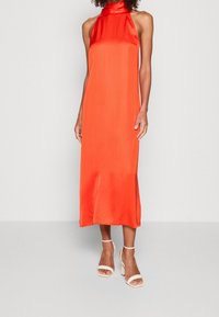 Frau in einem ärmellosen, hellorangefarbenen Satin-Midikleid mit hohem Ausschnitt und weißen, knöchelhohen Sandalen mit Absatz auf einem hellen Hintergrund.