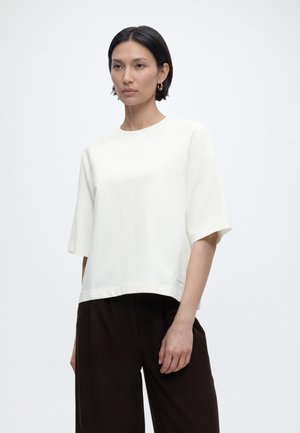 Γυναίκα με κοντά σκούρα μαλλιά, που φοράει χρυσούς κρίκους στα αυτιά, λευκό χαλαρό τοπ με κοντά μανίκια και σκούρο, φαρδύ παντελόνι, στέκεται μπροστά σε απλό φόντο.