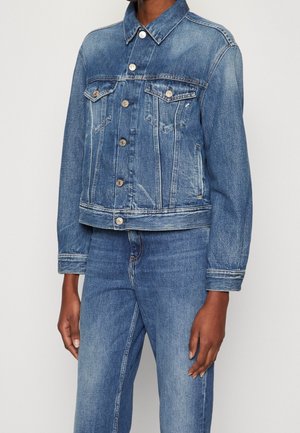 Personne portant une veste en jean bleu délavé moyen avec des boutons argentés et un jean en jean bleu assorti, debout devant un fond uni clair.