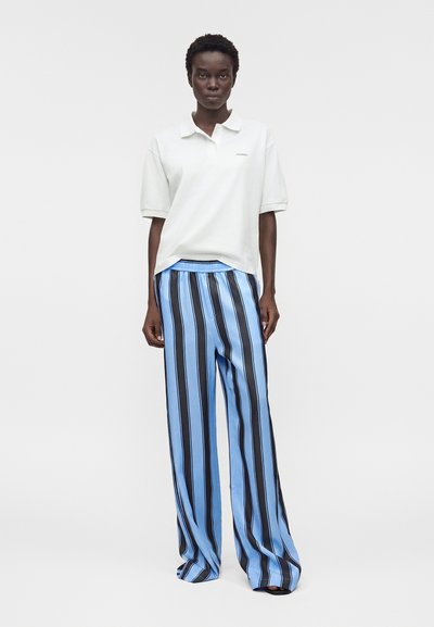 Άτομο που στέκεται φορώντας λευκό πόλο με κοντά μανίκια και φαρδιά μπλε παντελόνια με μαύρες κάθετες ρίγες, σε μονόχρωμο φόντο.