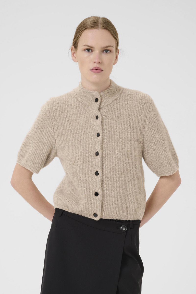 Beige strikket kortermet cardigan med høy krage, med svarte knapper foran og et teksturert stoff. Kombinert med svarte bukser.