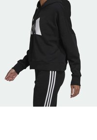 Felpa nera corta con logo bianco, dotata di cappuccio con cordino e polsini a costine. Abbinata a pantaloni neri con tre strisce bianche.