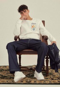 Sweatshirt blanc avec motif floral, associé à un jean bleu foncé. Modèle assis sur une chaise sur un tapis à motifs, portant des baskets blanches.