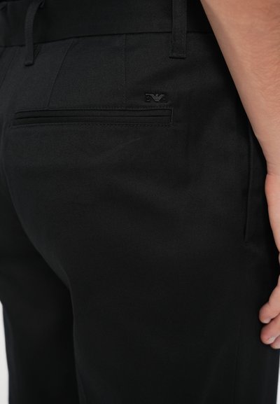 Čierne nohavice s hladkou textúrou, vybavené zadným vreckom a jemným detailom loga. Strih na mieru s opaskovými očkami a hladkým dizajnom.