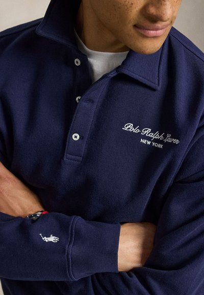 Άτομο που φοράει μπλε σκούρο πουκάμισο Polo Ralph Lauren με κεντημένο λευκό λογότυπο στο στήθος και στο μανίκι, με σταυρωμένα χέρια.