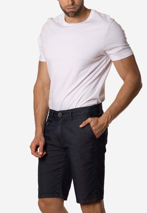 Hombre vistiendo una camiseta blanca de manga corta y unos pantalones cortos oscuros hasta la rodilla, de pie con una mano en el bolsillo.