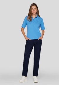 Hellblaues Poloshirt mit kurzen Ärmeln, zwei Knöpfen und einem kleinen Logo. Kombiniert mit dunkelblauen Hosen und weißen Turnschuhen.