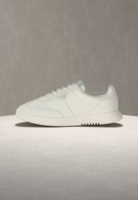 Bílá nízká teniska se šněrováním vpředu, decentním logem na boku a silnou gumovou podrážkou, zobrazená na neutrálním povrchu a pozadí.