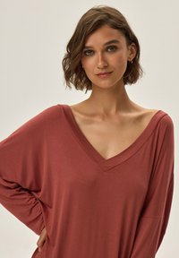 Rustowy, oversize top z dekoltem w serek, wykonany z miękkiego materiału, z szerokimi rękawami i luźnym krojem, o gładkiej teksturze i minimalnym zdobieniu.