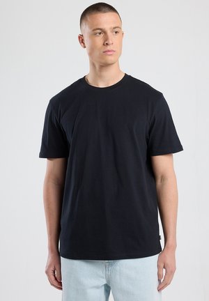 Zwart T-shirt met korte mouwen van katoen. Ronde halsontwerp, relaxte pasvorm en minimale stiksels. Draagt met lichtblauwe jeans.