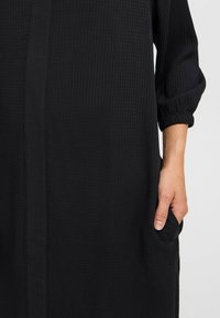 Fekete ruha texturált anyaggal, rugalmas mandzsettákkal és oldalsó zsebekkel, laza illeszkedéssel és közepes hosszúságú siluettel.