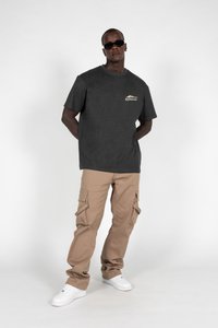 Tricou gri cu guler rotund și logo, asortat cu pantaloni cargo bej cu multiple buzunare și adidași albi. Model purtând ochelari de soare negri.