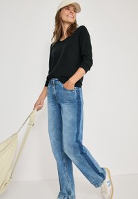 Hellblaue Jeans mit seitlichem Streifen, kombiniert mit einem schwarzen Pullover und einer beigen Mütze. Beiger Tasche und weißen Sneakers mit blauen Akzenten.