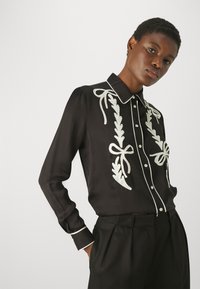 Blusa nera a maniche lunghe con design floreale ricamato bianco e fiocchi, dotata di colletto e chiusura frontale con bottoni. Tessuto dalla texture morbida.