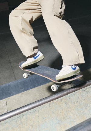 Personne portant un pantalon beige et des baskets blanches avec des détails bleus réalisant une figure de skateboard sur une rampe.