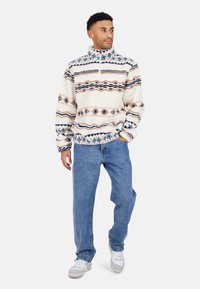Uomo che indossa un pullover in pile crema con motivo, jeans blu e sneakers bianche, in piedi con una mano in tasca, guardando a destra.