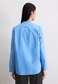 Himmelblaues, langärmliges Hemd mit geradem Schnitt, geknöpften Manschetten und abgerundetem Saum. Glattes Material, minimale Details auf der Rückseite.