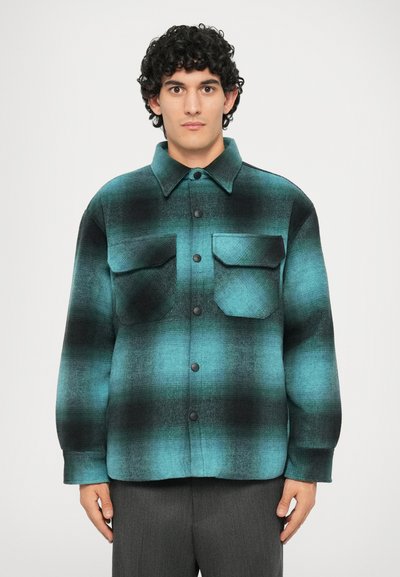 Camicia di flanella in fantasia a quadri teal e nera, con colletto, chiusura a bottoni e due tasche sul petto. Texture morbida e vestibilità rilassata.