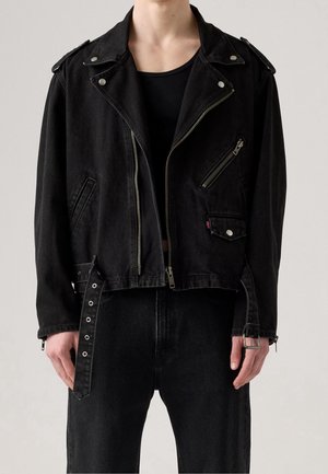 Personne portant une veste courte de motard noire avec des fermetures éclair argentées, des boutons-pression et des bretelles réglables, par-dessus un pantalon noir et un haut noir.